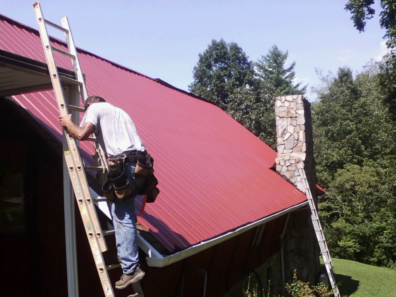 Roofer on a ladder beside a red metal roof and stone chimney — Soffit & Fascia Repair by Roofing Brothers in Old Town